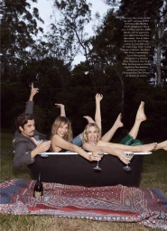 Margot Robbie and friends posing playfully in an outdoor black bathtub on a patterned rug with drink glasses at twilight.
