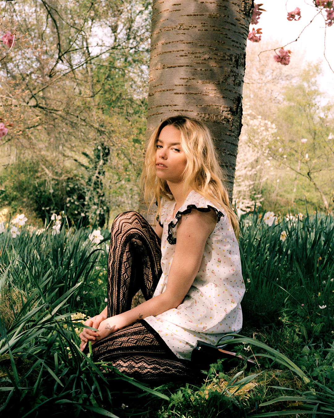 A blonde woman in a patterned dress and tights sits near a tree on grass surrounded by white and green plants and cherry blossoms.