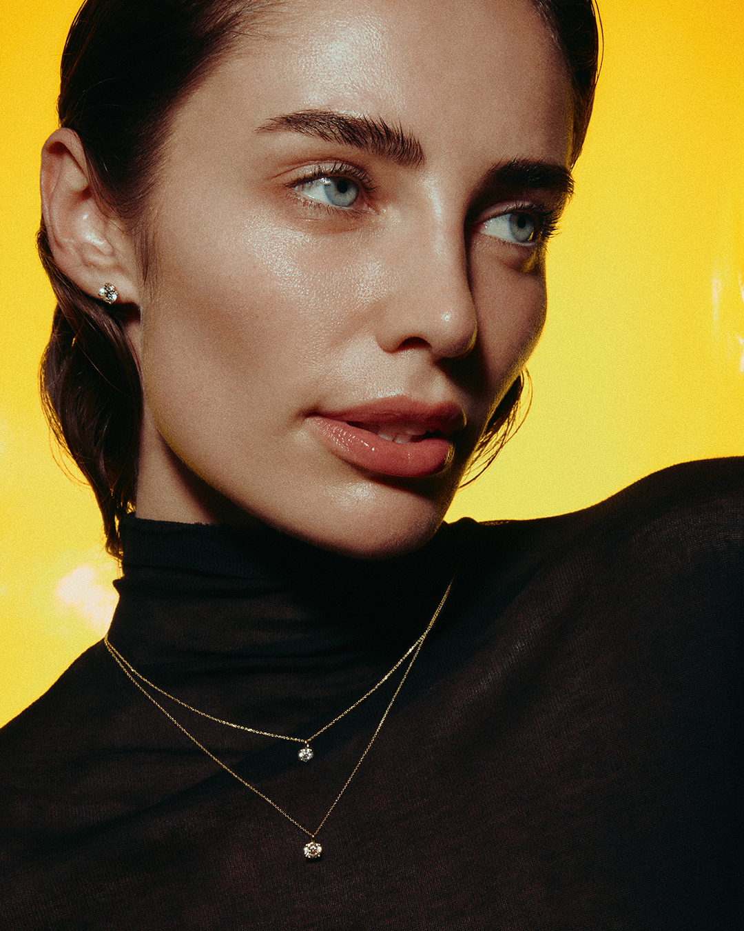 Close-up portrait of a woman with light skin and blue eyes wearing a black top and layered gold necklaces against a yellow background.