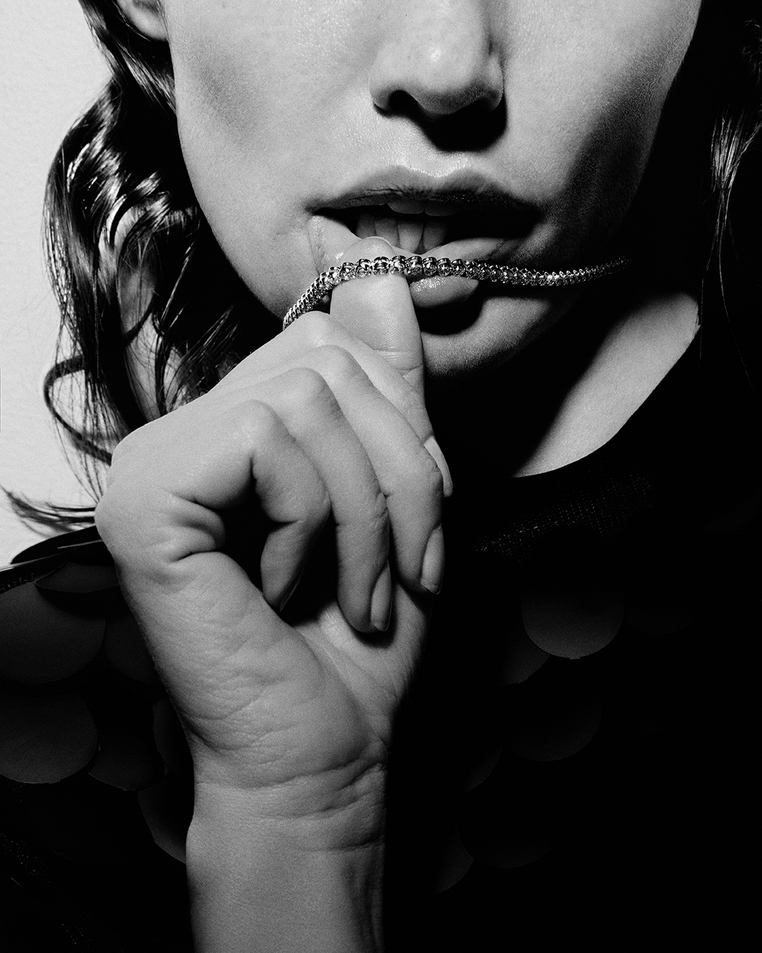 Close-up black and white image of a woman holding a sparkling necklace in her hand and biting it with her lips, set against a bright backdrop.