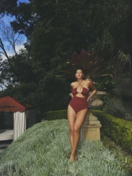 Model posing in a burgundy cutout halter swimsuit next to a large stone garden urn surrounded by lush foliage.