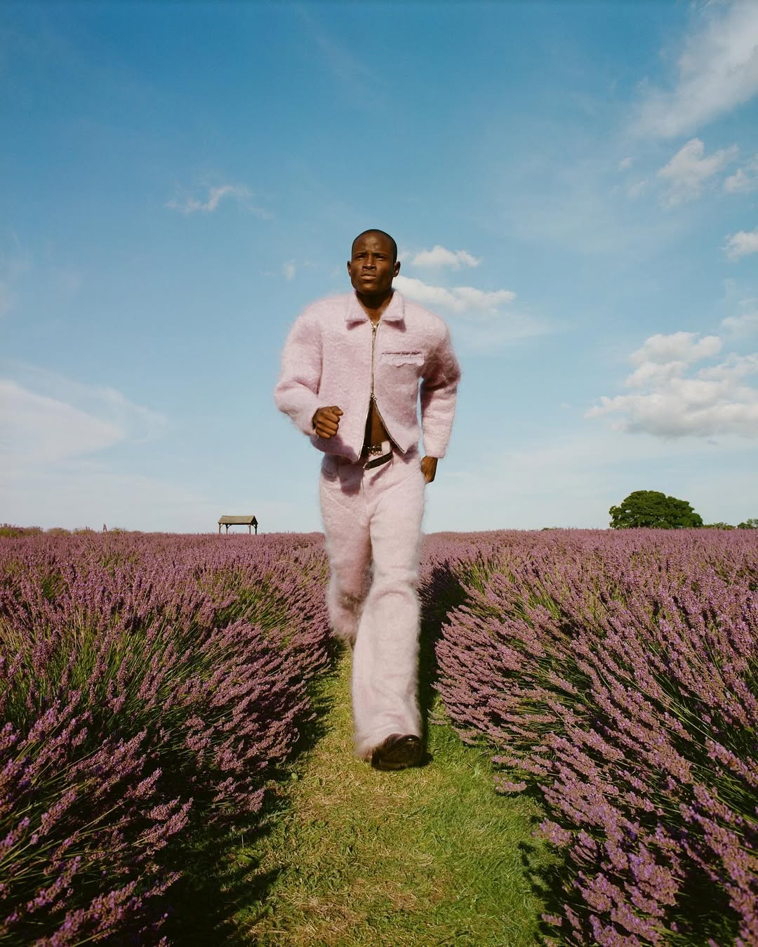 Dark-skinned man running through field of purple lavender wearing pink jacket and pants under a blue sky with light clouds.