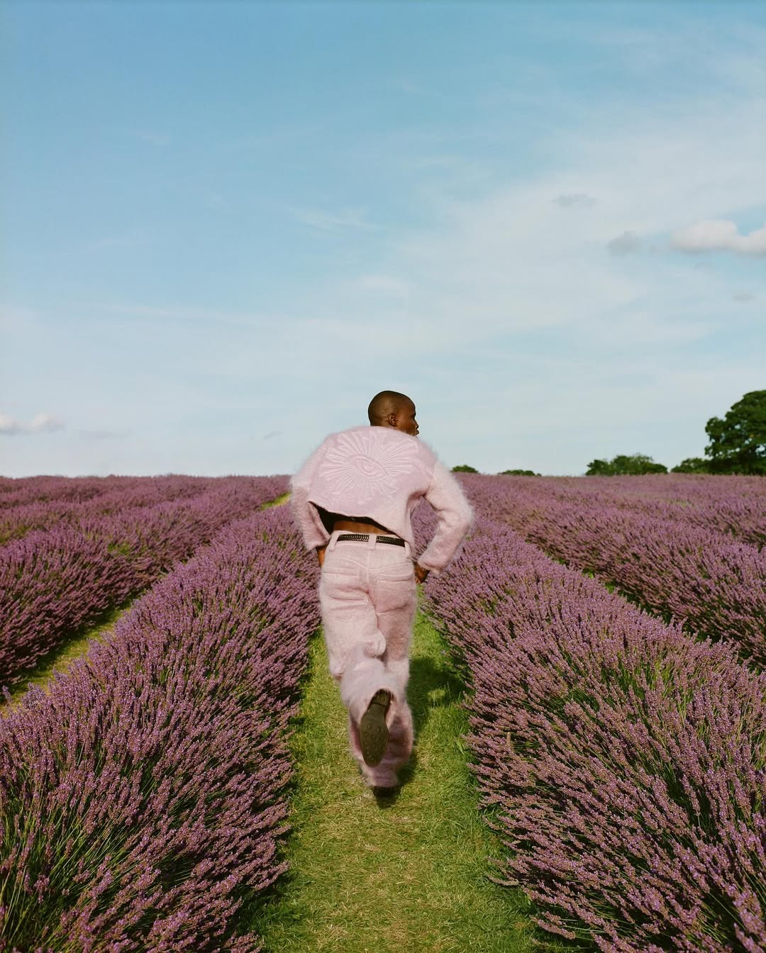 A man runs through a lavender field under a blue sky, wearing a light pink jacket and pants, captured in a medium shot.