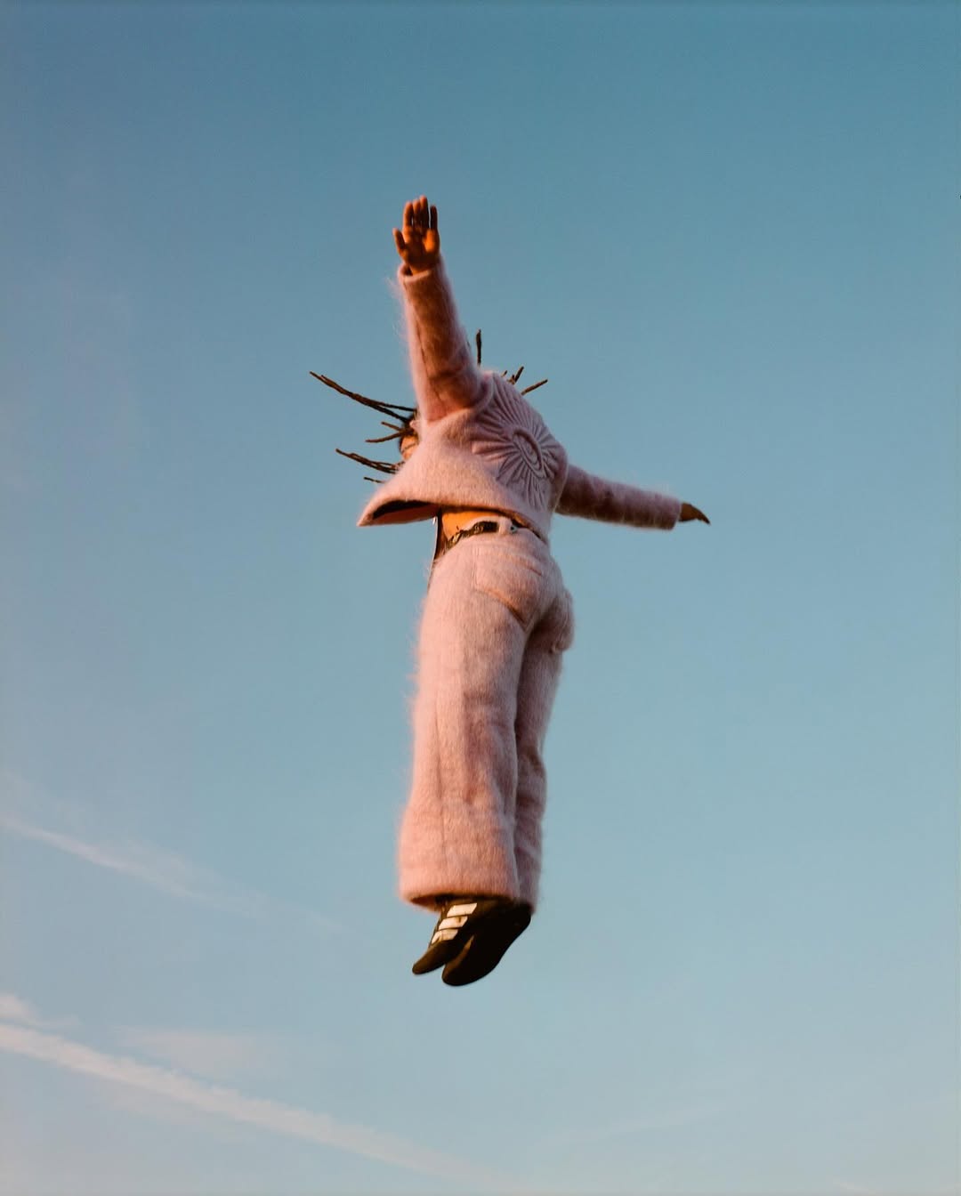 Fashionable person levitates against a clear blue sky, wearing a furry pink jacket and pants with distinctive black and white shoes.