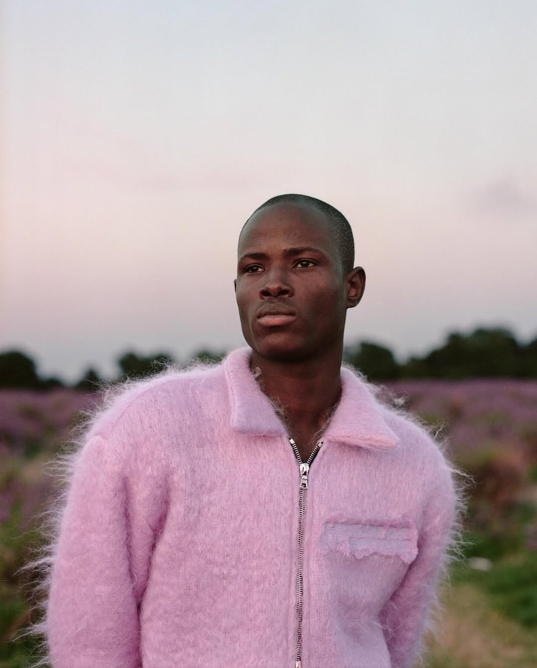 Portrait of a dark-skinned man in a pink jacket in a field with purple flowers. He's looking off to the side with a neutral expression.