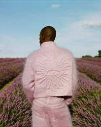 Man wearing a fluffy pink textured jacket and matching trousers standing amidst a vast lavender field under a clear blue sky