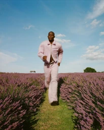 Black man running through a blooming lavender field wearing a fuzzy pink jacket and matching trousers under a clear blue sky