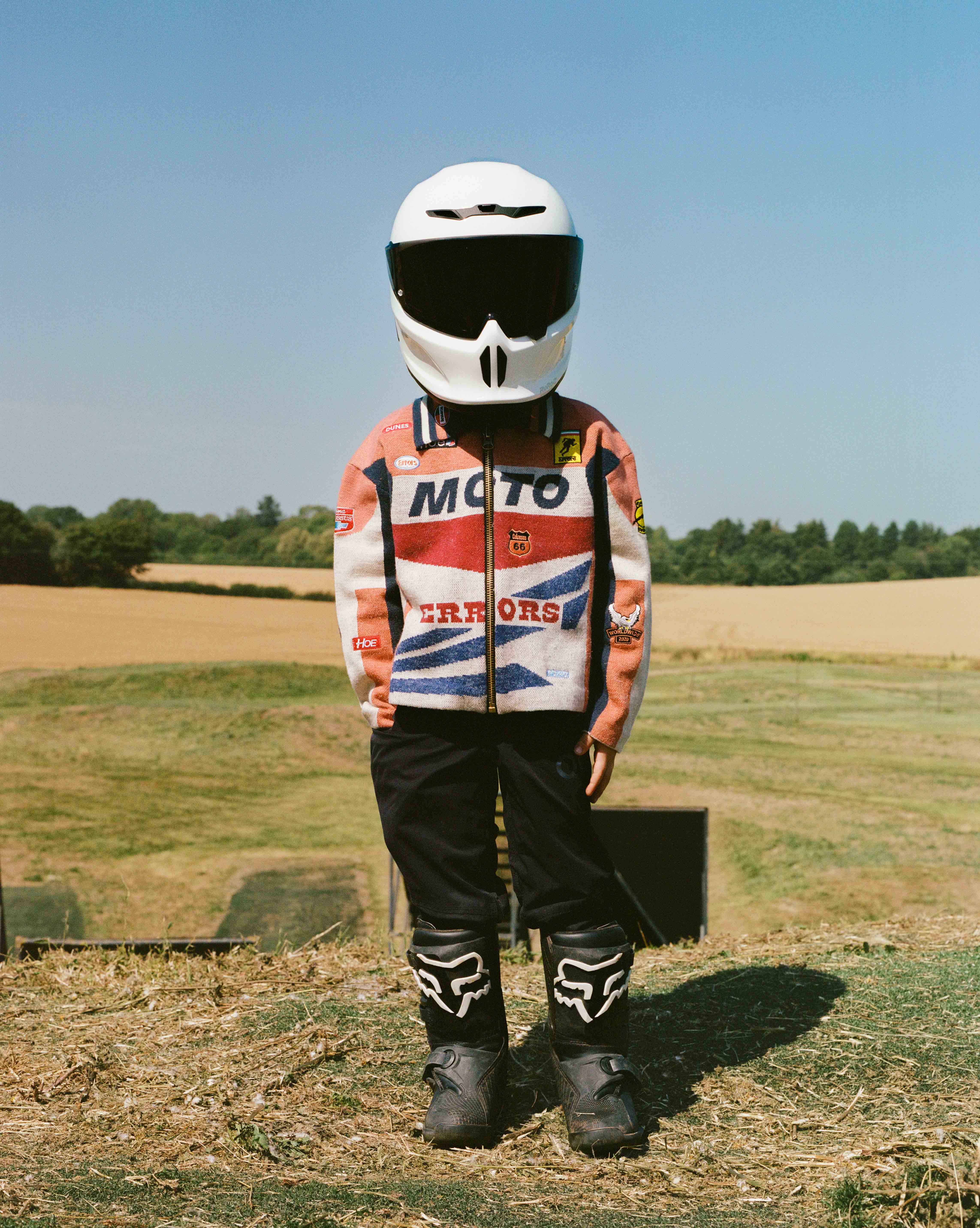 Child in helmet and racing jacket stands in field on sunny day. Black pants, Fox-brand boots complete the look. Trees and blue sky.