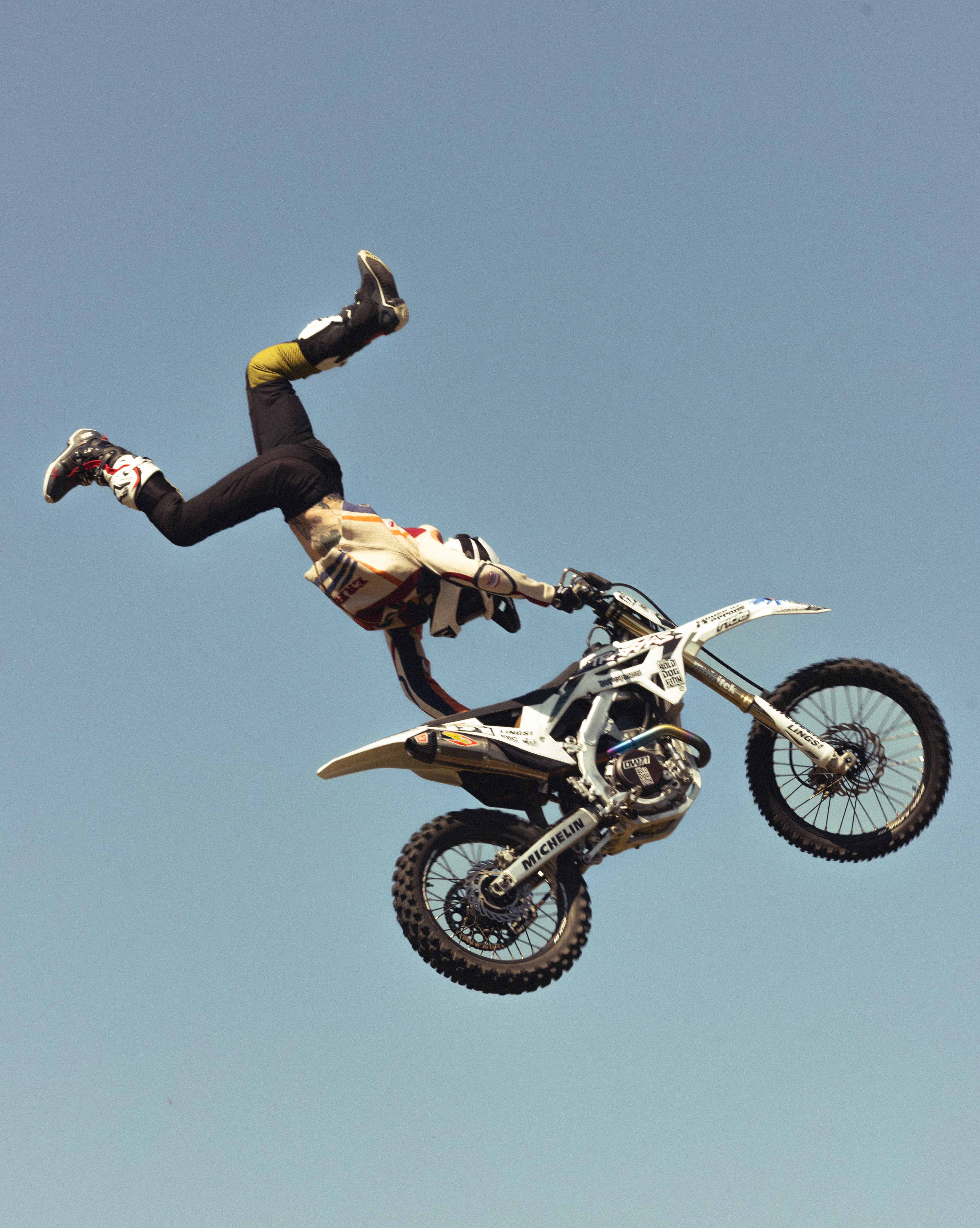 A motorcyclist performs a trick in the air on a motocross bike against a clear blue sky, showcasing action and skill in an outdoor setting.