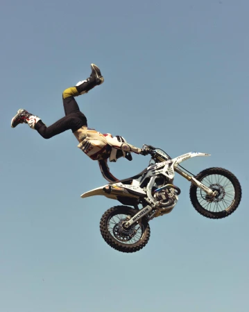 A motorcyclist performs a trick in the air on a motocross bike against a clear blue sky, showcasing action and skill in an outdoor setting.