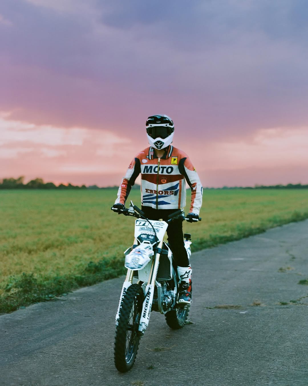 Person riding a white and blue dirt bike on an asphalt road, with green fields and a vibrant sky in the background.