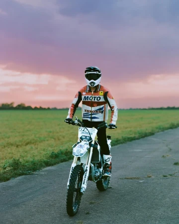Person riding a white and blue dirt bike on an asphalt road, with green fields and a vibrant sky in the background.
