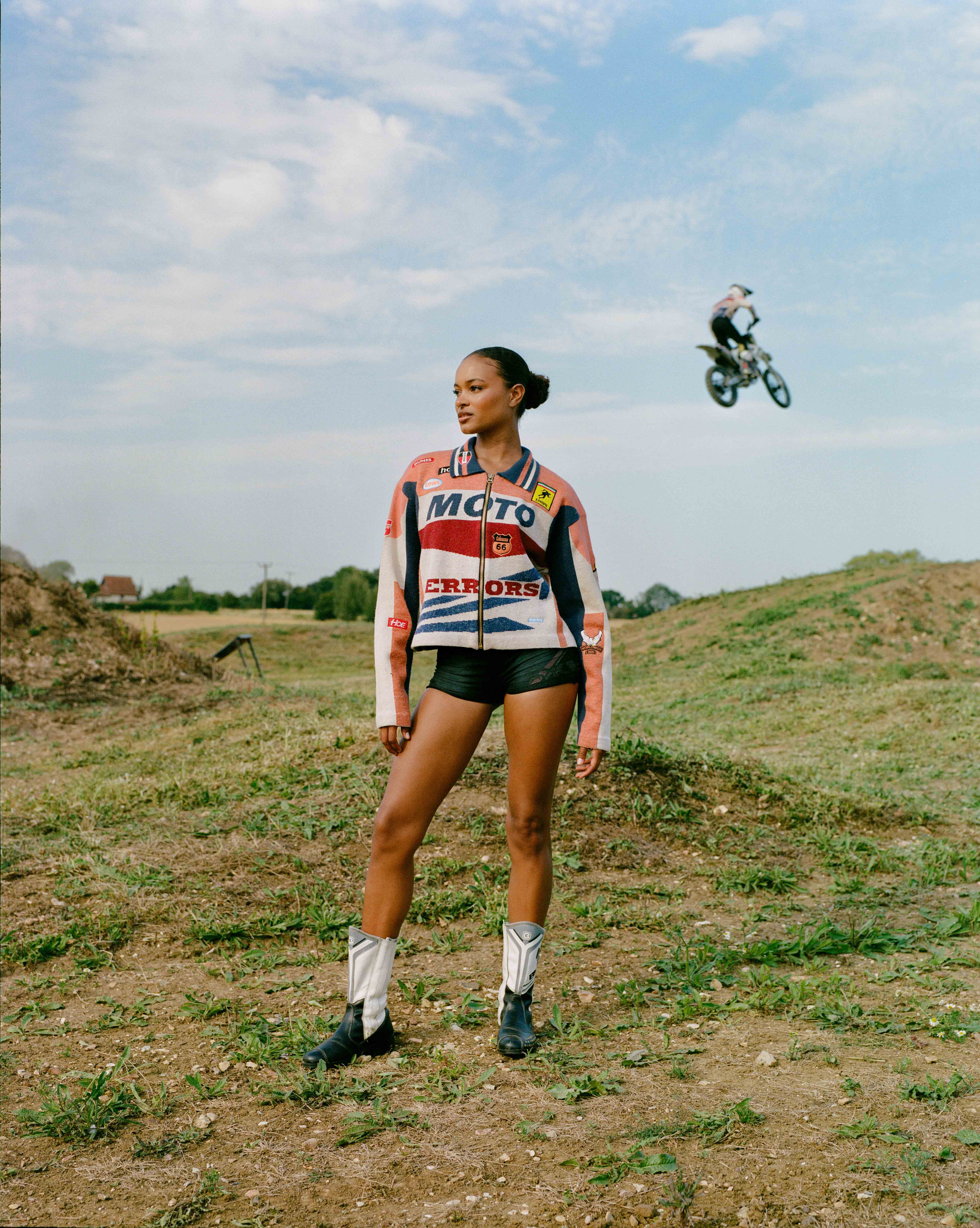 A woman in a racing jacket and cowboy boots stands in a field with a motocross rider jumping in the background on a sunny day.