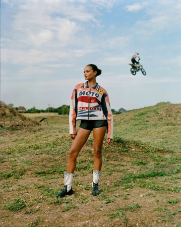 A woman in a racing jacket and cowboy boots stands in a field with a motocross rider jumping in the background on a sunny day.