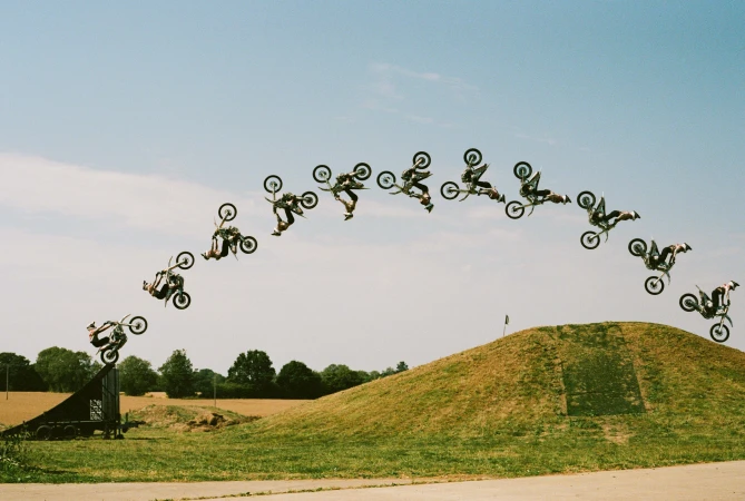 Motocross rider performs a flip on a dirt bike multiple times in a sequence, captured outdoors with a ramp and a clear blue sky background.