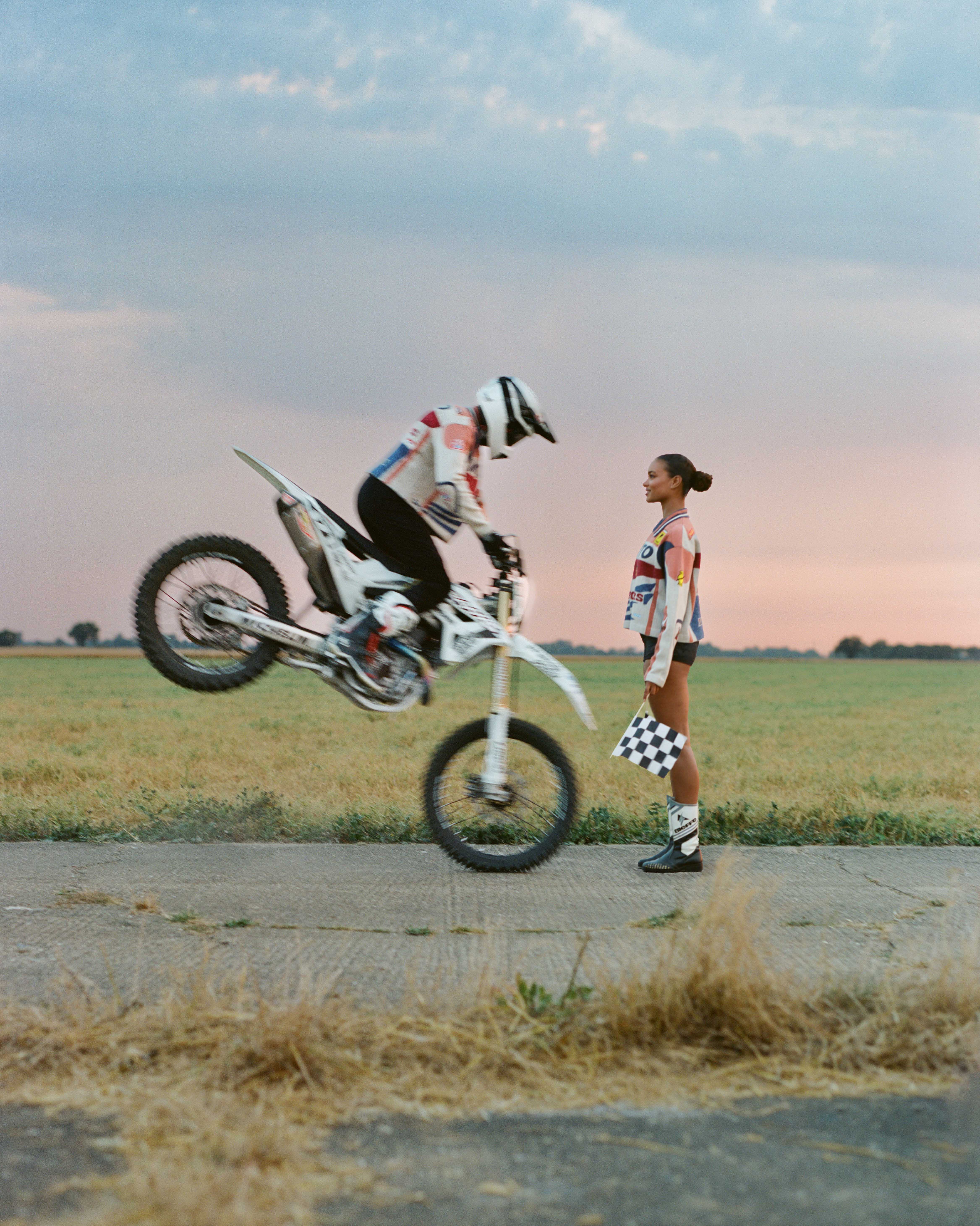 Dirt bike rider performs wheelie on a rural road, with a woman holding a checkered flag nearby at sunset. High-speed action shot.