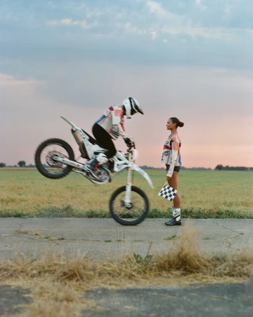 Dirt bike rider performs wheelie on a rural road, with a woman holding a checkered flag nearby at sunset. High-speed action shot.