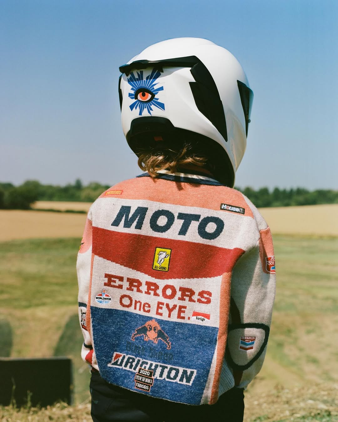 Child in motorcycle helmet and racing jacket stands in a grassy field under a clear blue sky on a sunny day, captured in a medium shot.