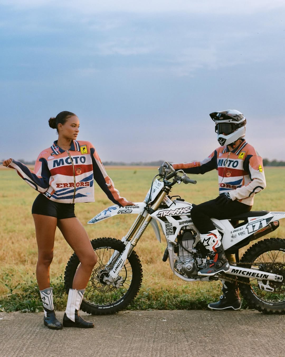 Two people pose next to a white dirt bike on a rural road under a cloudy sky, wearing matching racing-style jackets and boots.