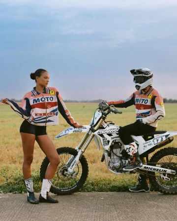 Two people pose next to a white dirt bike on a rural road under a cloudy sky, wearing matching racing-style jackets and boots.