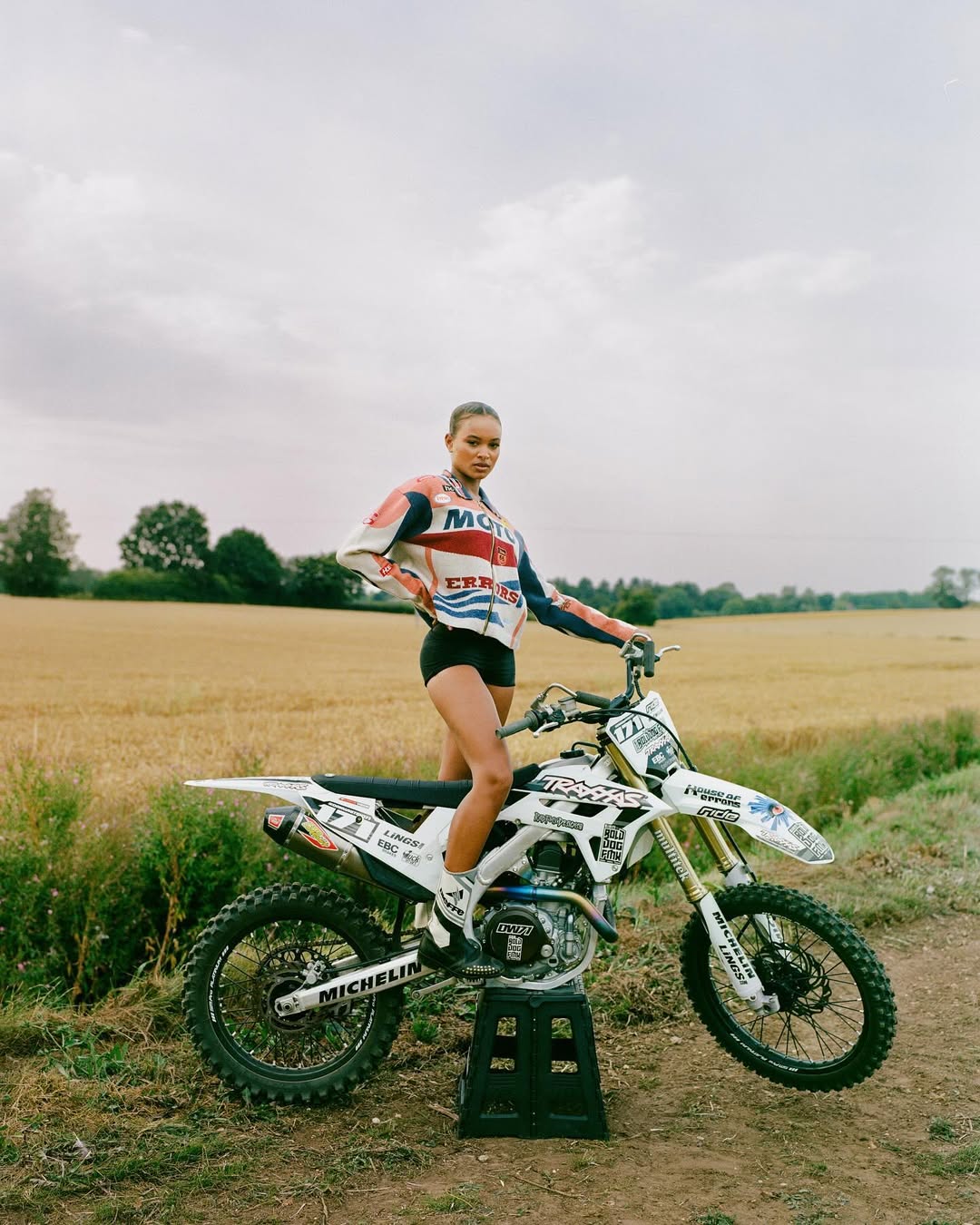 A woman in a racing suit poses with a motocross motorcycle on a stand in a grassy field under a cloudy sky in a medium outdoor shot.