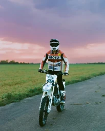 Person wearing a helmet and leather jacket riding a dirt bike on an asphalt road through an open field under a cloudy sunset sky.