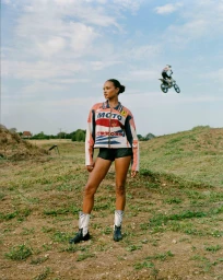 Model wearing a Moto Errors jacket and motocross boots standing outdoors on a dirt hill with a jumping dirt bike in the background