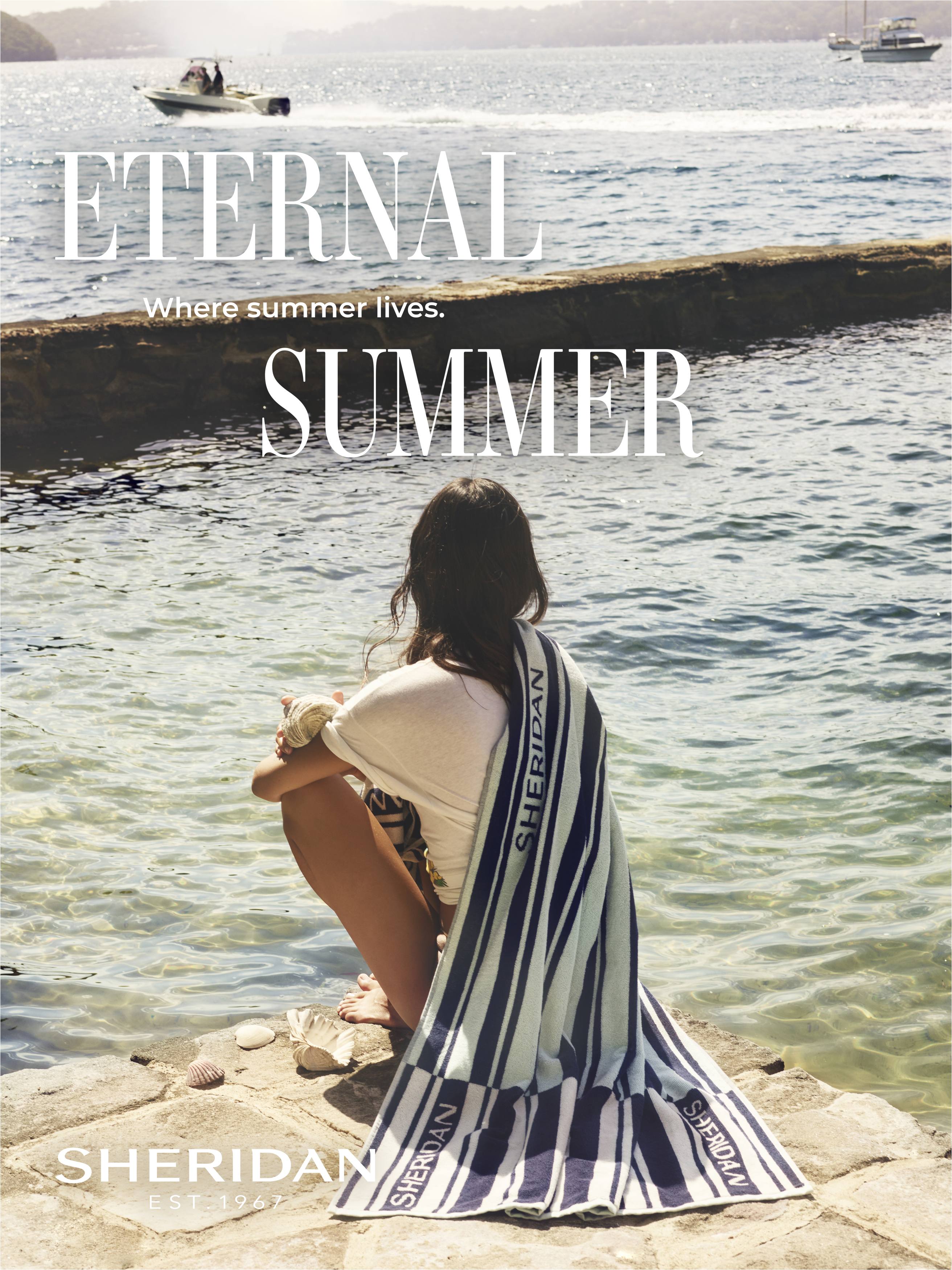 Woman sits on a rock wall by the ocean, draped in a Sheridan towel with a motorboat in the distance, capturing a serene summer scene.