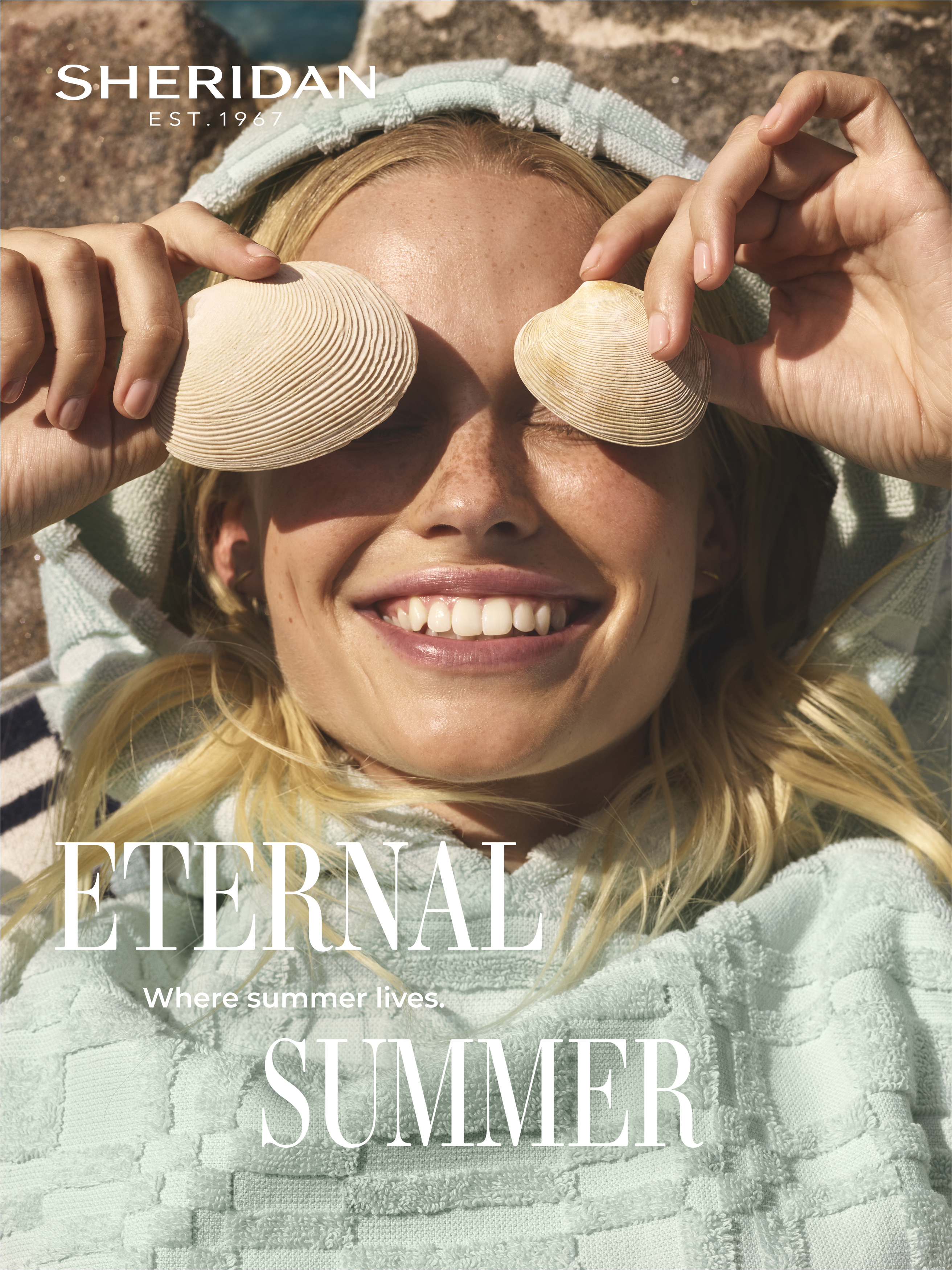 Smiling blonde woman in a hooded towel holds seashells over her eyes, bathed in soft sunlight. Sheridan brand logo visible.