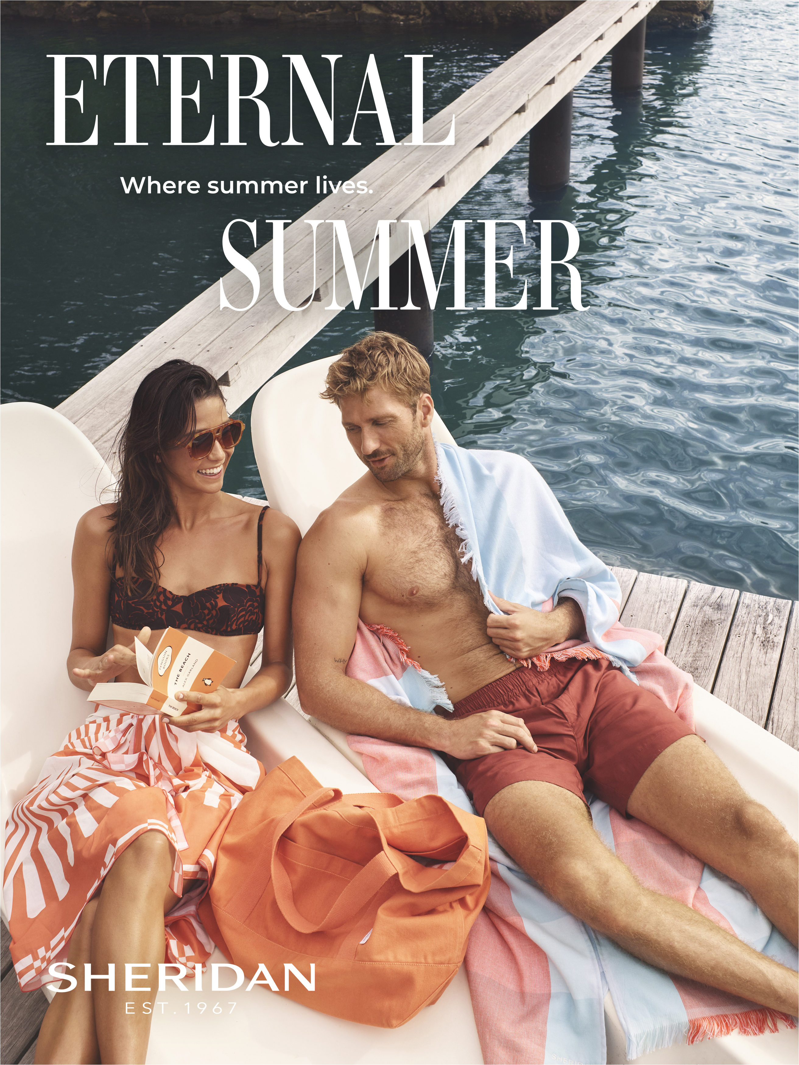 Two adults lounge near the water's edge, one holding a product, both covered in colorful towels, with a matching orange tote bag.