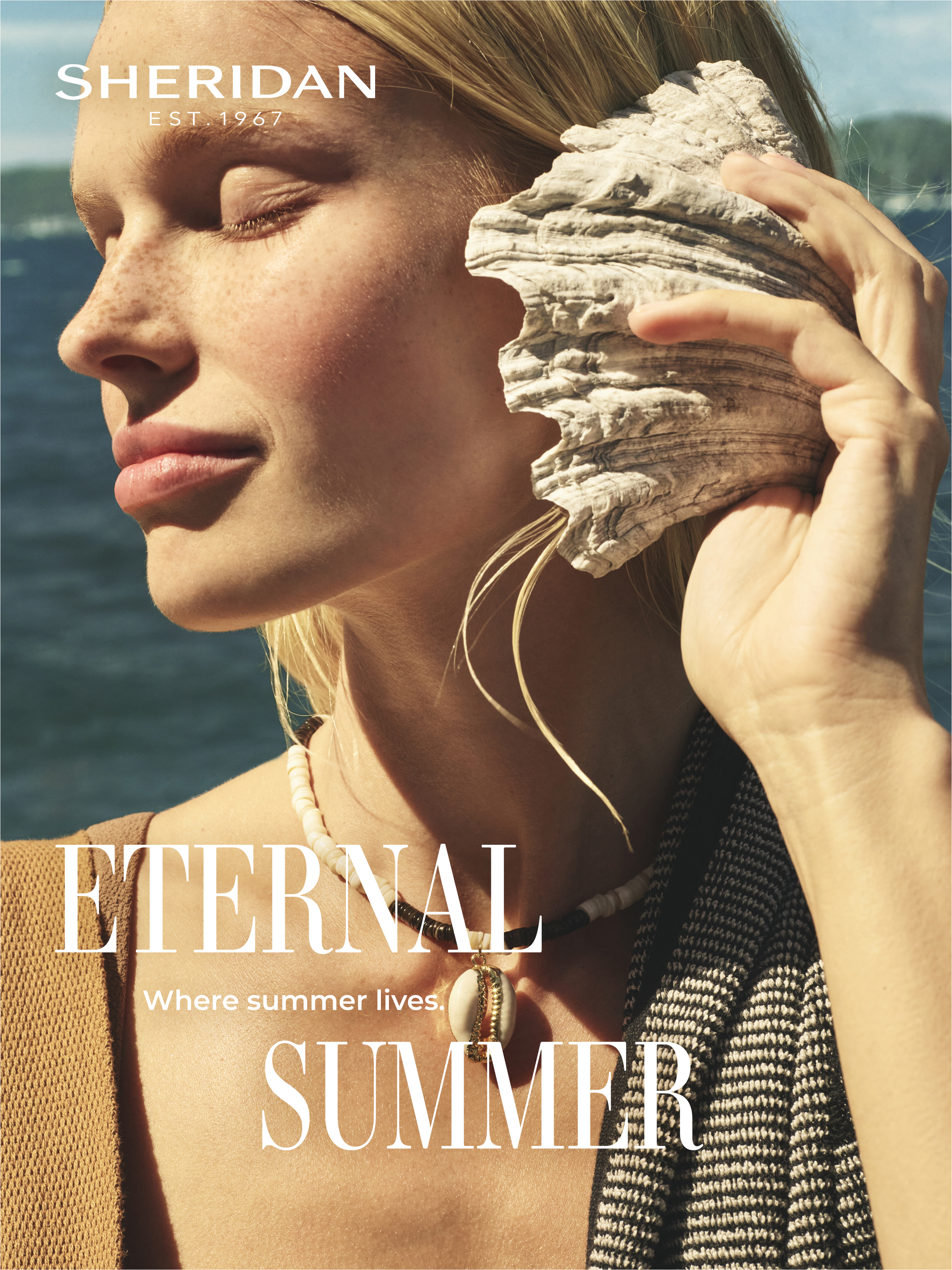A woman with fair skin and blonde hair holds a seashell to her ear in front of a blue ocean. Warm, natural lighting.