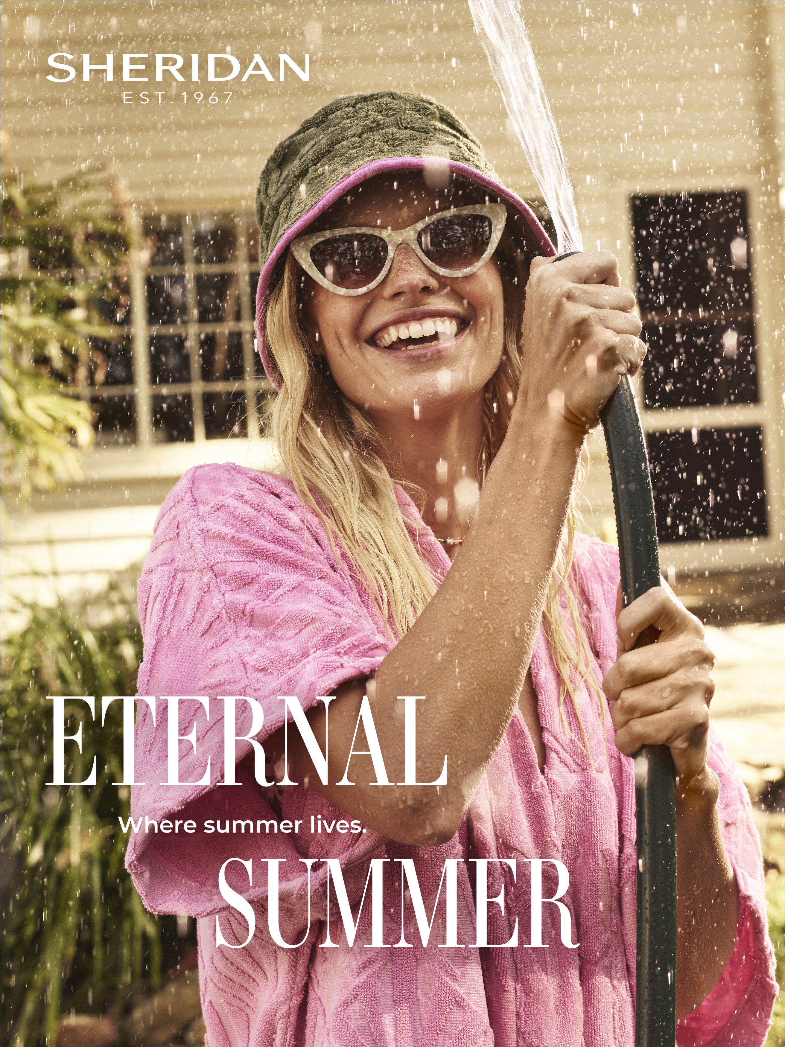 A cheerful woman in sunglasses and a pink robe smiles while holding a garden hose, enjoying a sunny day in a lush outdoor setting.