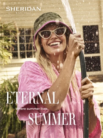 A cheerful woman in sunglasses and a pink robe smiles while holding a garden hose, enjoying a sunny day in a lush outdoor setting.