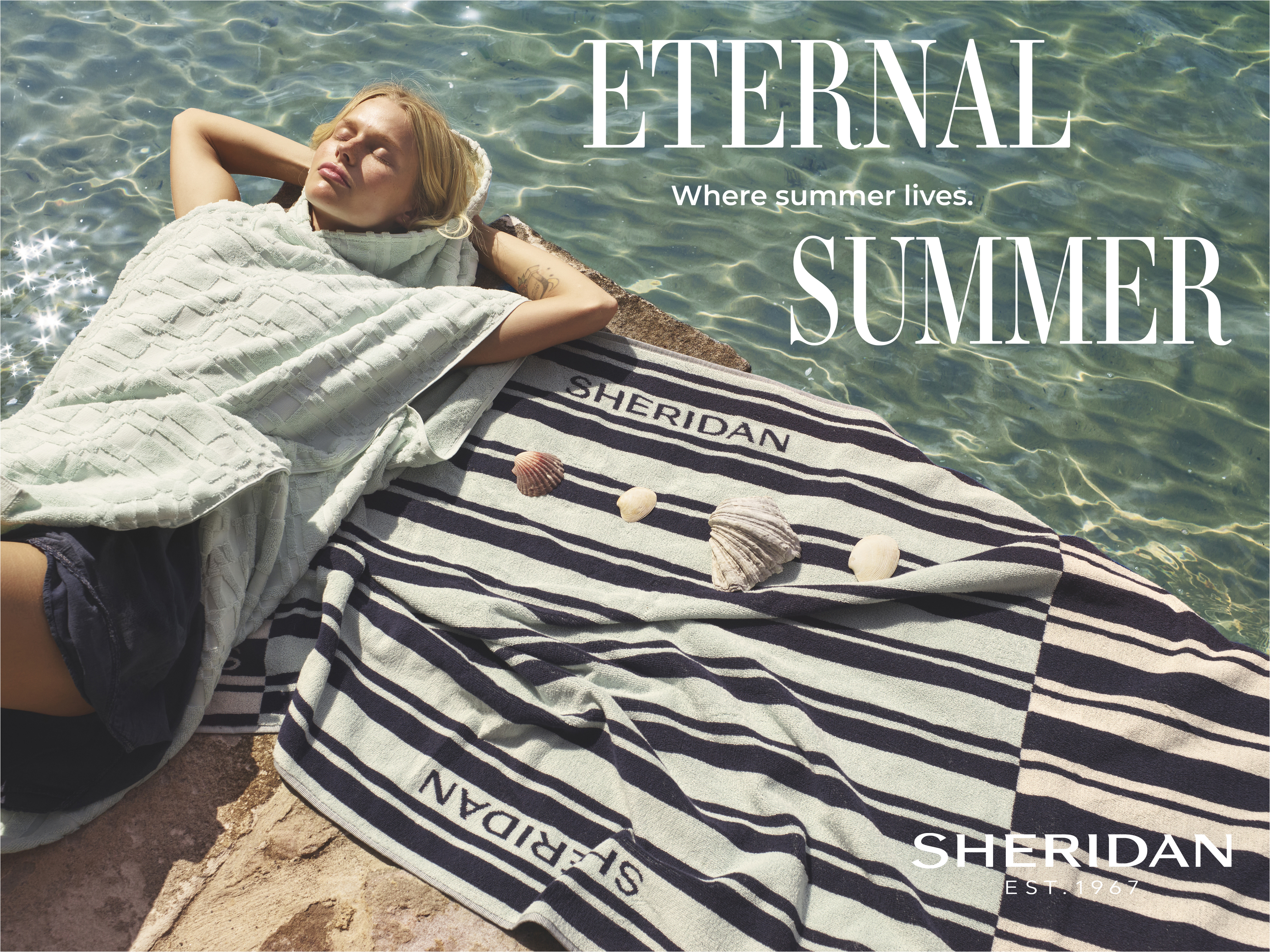 Relaxing summer scene: fair-skinned woman in cover-up on rock with striped towel and seashells by clear water under bright sunlight.