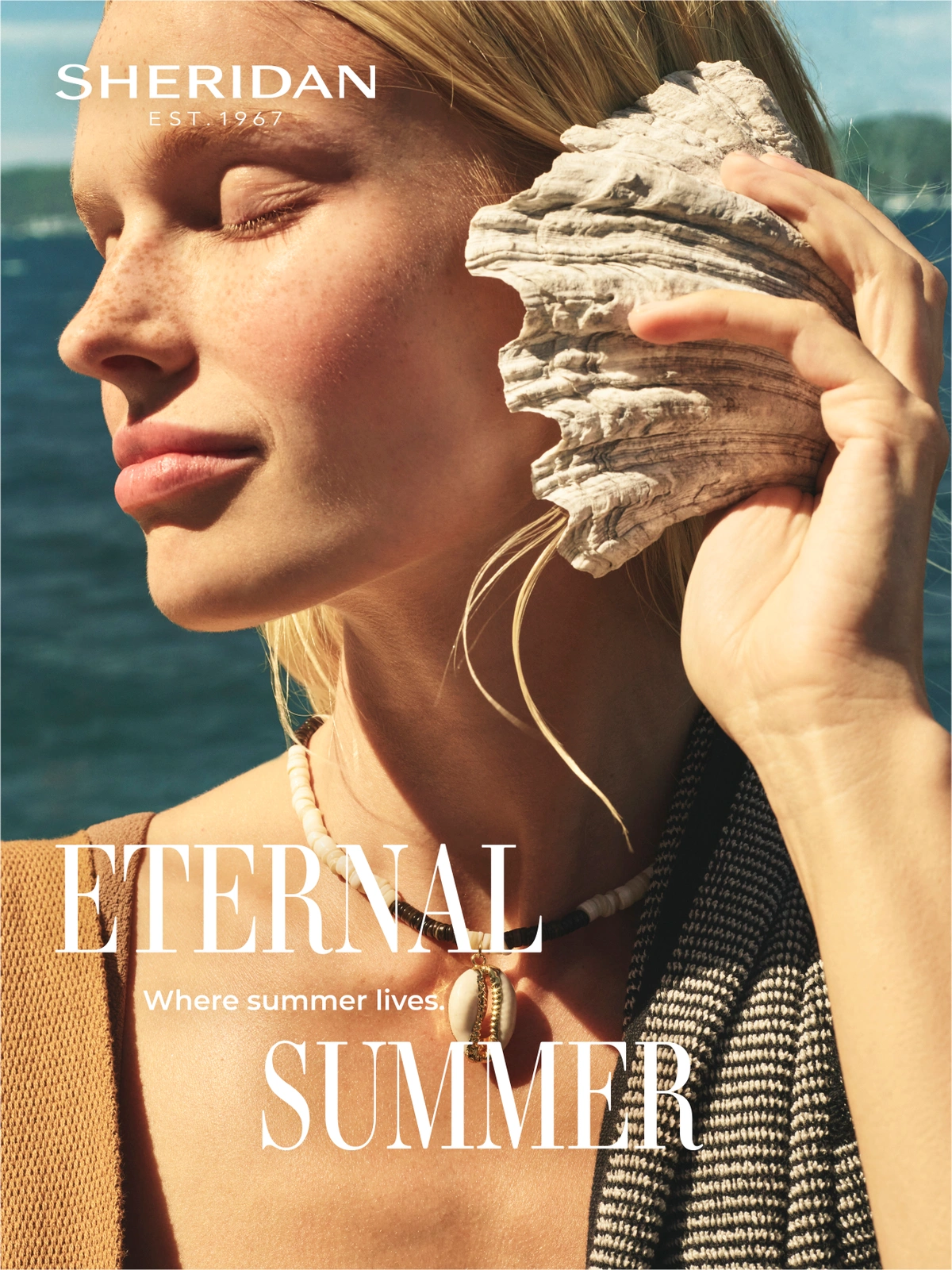 Close-up of a blonde woman holding a large seashell to her ear with the ocean visible in the background.
