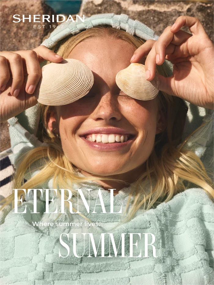 Close-up portrait of a smiling blonde woman holding seashells over her eyes, wearing a mint green hooded towel robe.