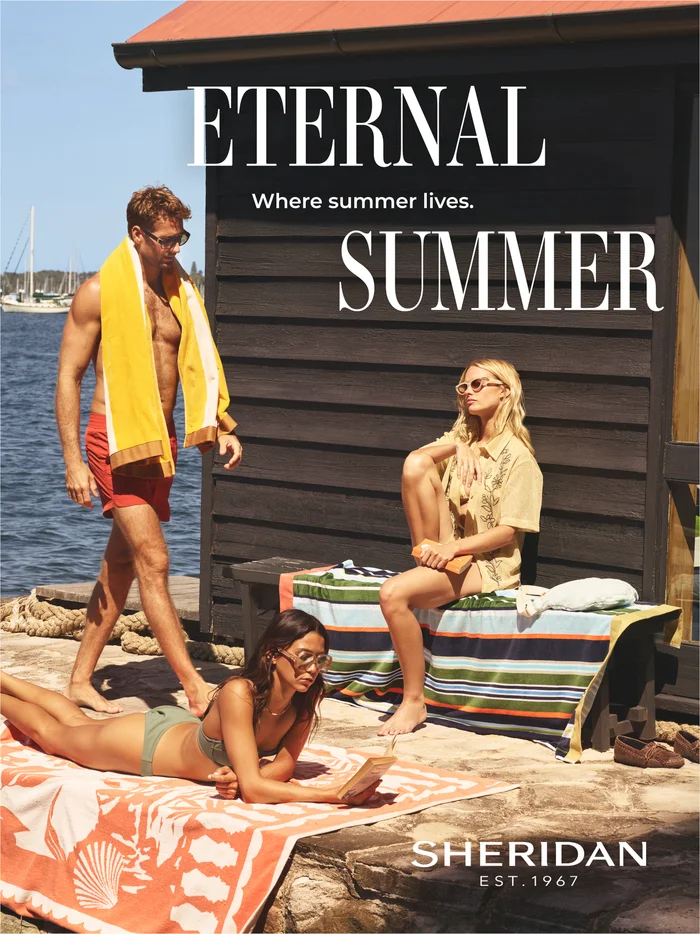 Three models relaxing outdoors by a dark wooden beach cabin with striped towels under bright sunlight