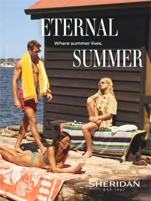 Three models relaxing outdoors by a dark wooden beach cabin with striped towels under bright sunlight