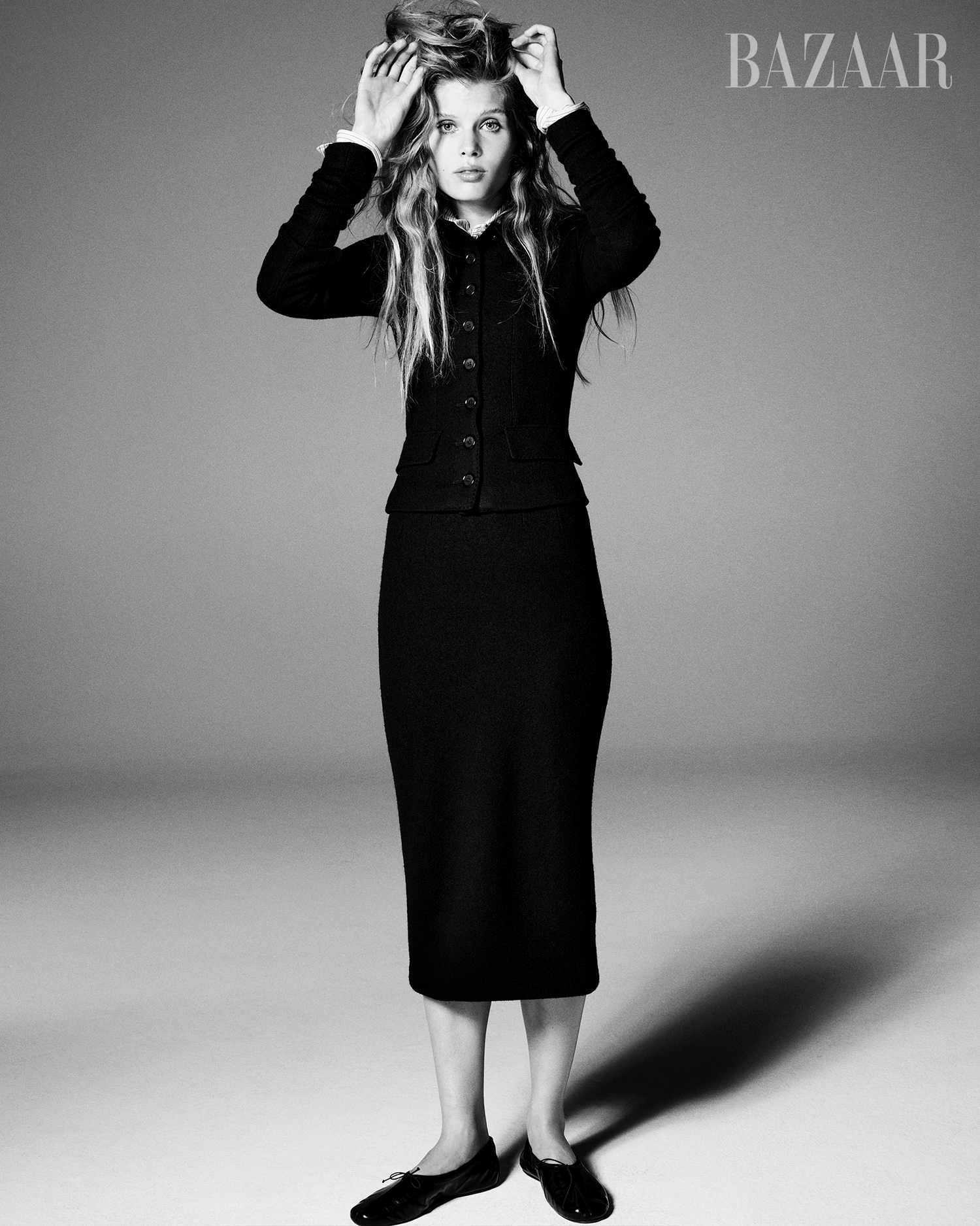 Model Ida Heiner adjusting hair in a black buttoned cardigan and midi skirt against a grey seamless studio background.