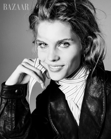 Black-and-white portrait of a smiling woman in a striped shirt and leather jacket against a grey backdrop; fashion-forward and engaging.