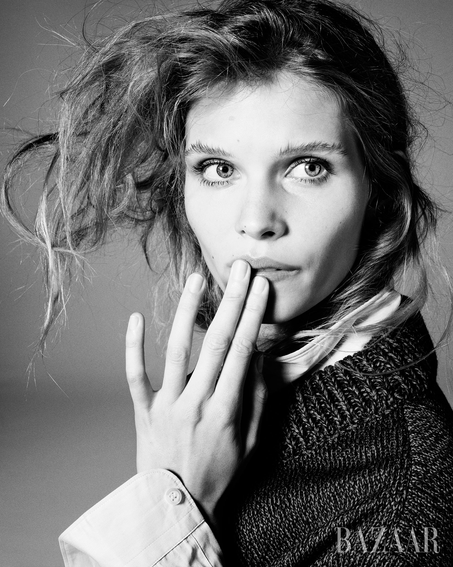Close-up black and white portrait of a young woman with tousled hair, looking at the camera with her hand near her mouth.