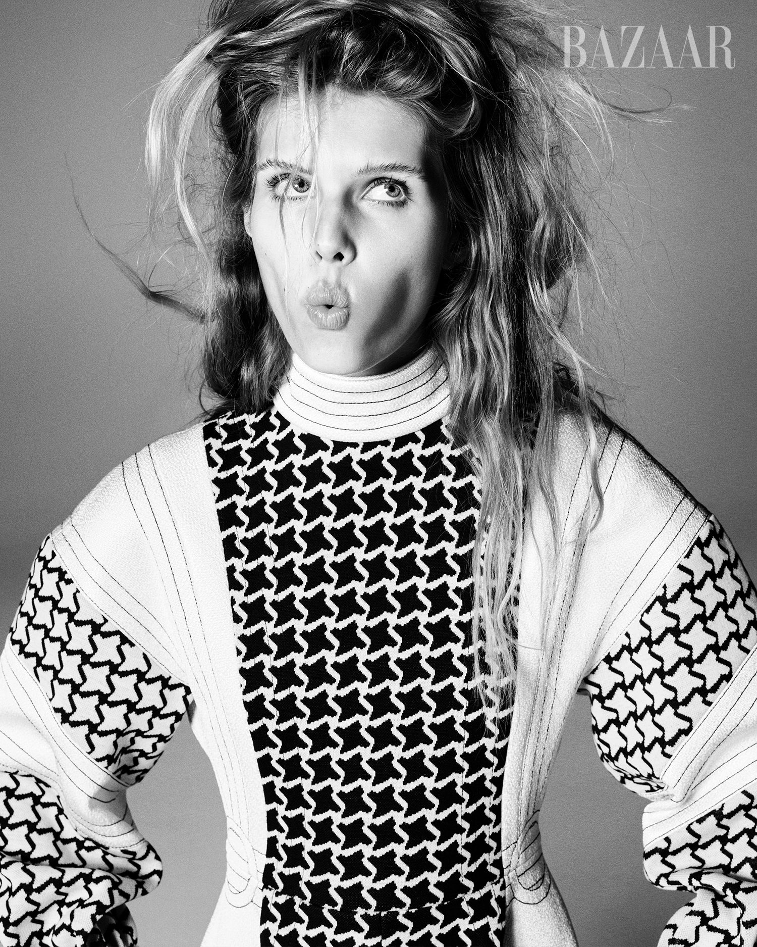 Black and white portrait of a young adult woman with long wavy hair, wearing a patterned top, and pulling a funny face in a studio.