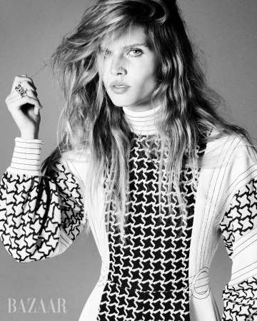 Black and white portrait of a stylish adult woman with long wavy hair, wearing a patterned dress, and rings against a grey backdrop.