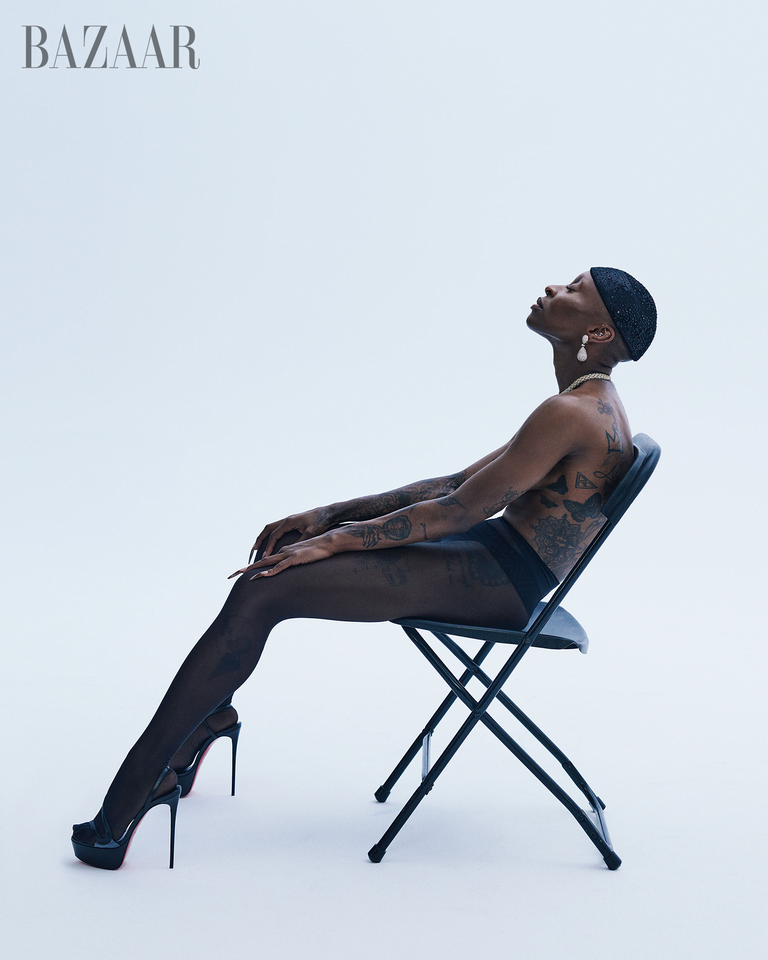 Profile of a black, bare-chested fashion model with arm tattoos wearing dark tights and high heels sitting on a black folding chair in a studio.