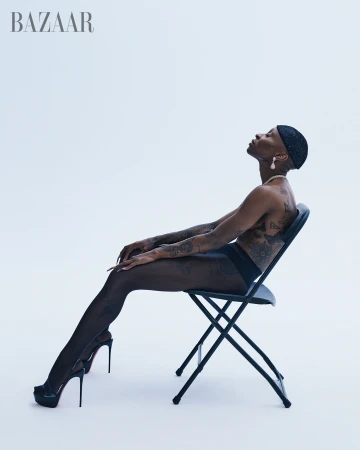 Profile of a black, bare-chested fashion model with arm tattoos wearing dark tights and high heels sitting on a black folding chair in a studio.