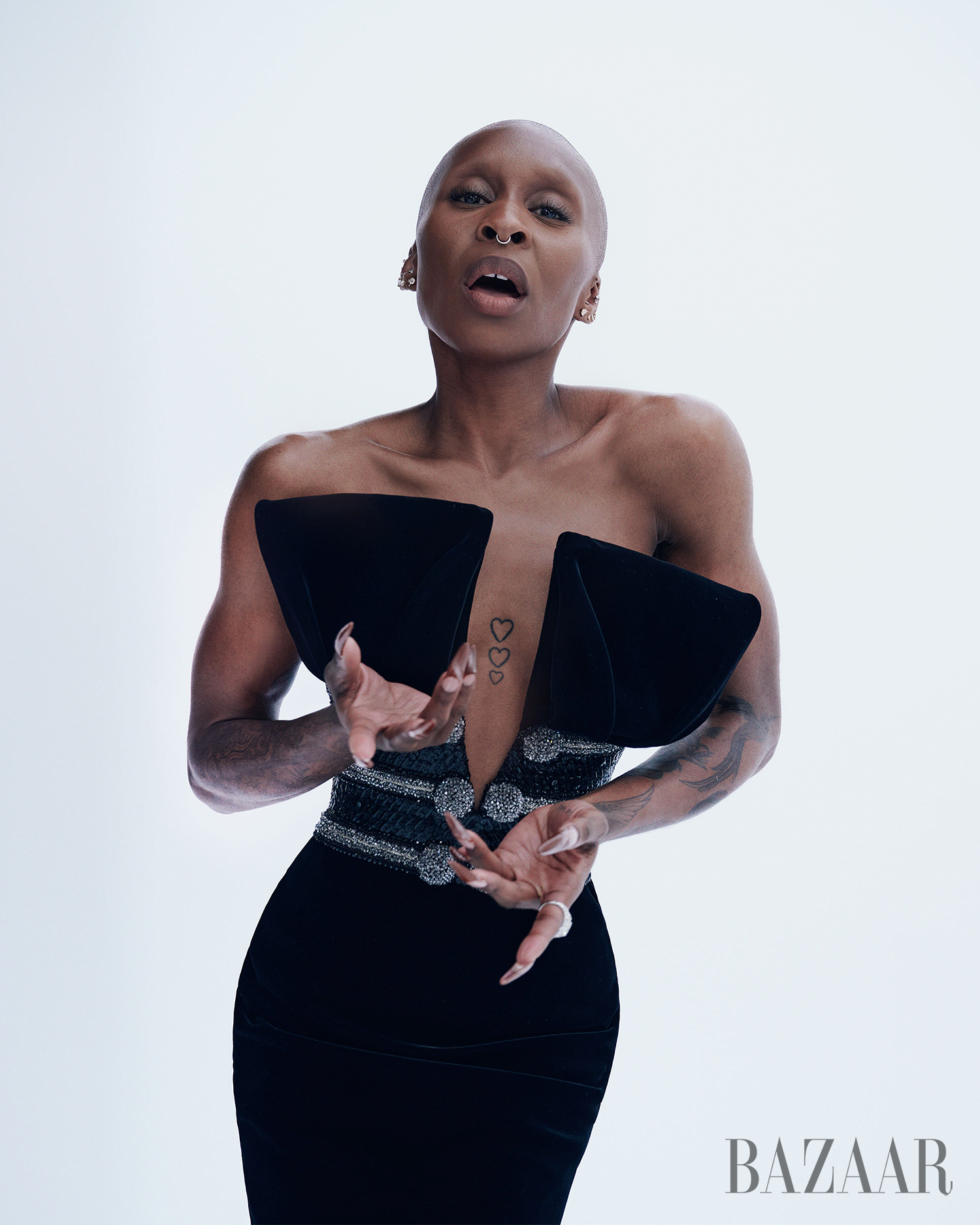 A slender, shaved-head Black woman poses in a studio wearing a black strapless dress with an embellished belt and pronounced shoulders.