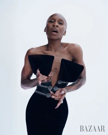 A slender, shaved-head Black woman poses in a studio wearing a black strapless dress with an embellished belt and pronounced shoulders.