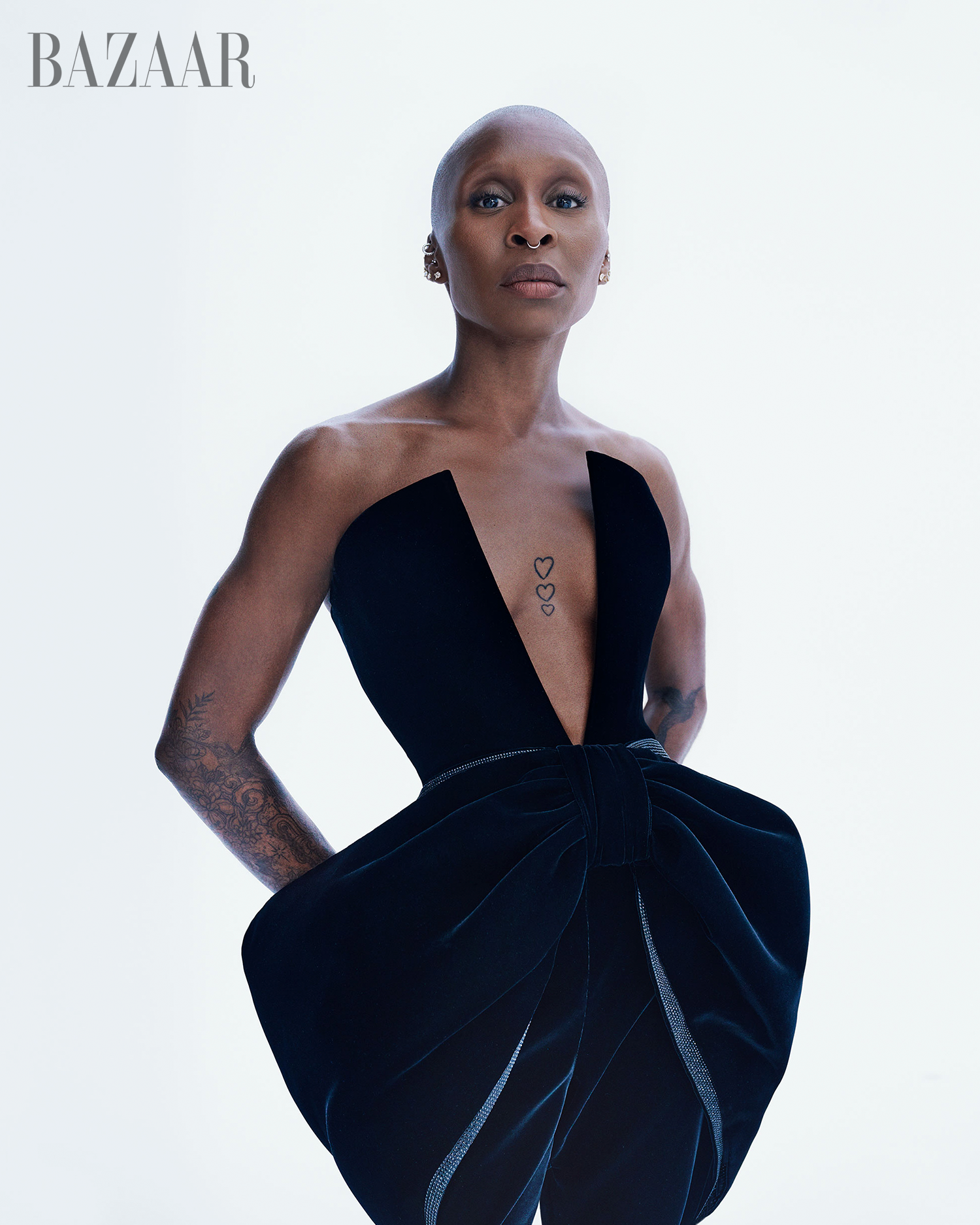 Close-up of a bald adult female wearing a black dress against a white background in a studio setting; fashion editorial shot. 