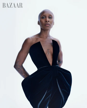 Close-up of a bald adult female wearing a black dress against a white background in a studio setting; fashion editorial shot. 
