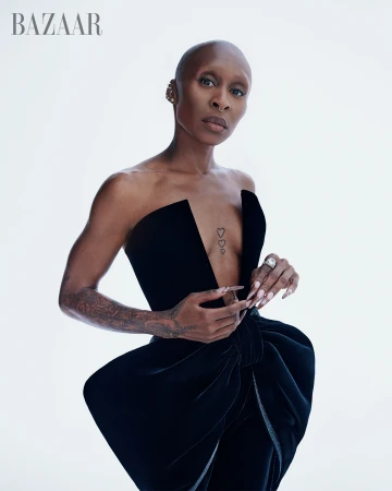 Striking portrait of a bald woman with visible tattoos wearing a black strapless dress with a large bow against a white backdrop.